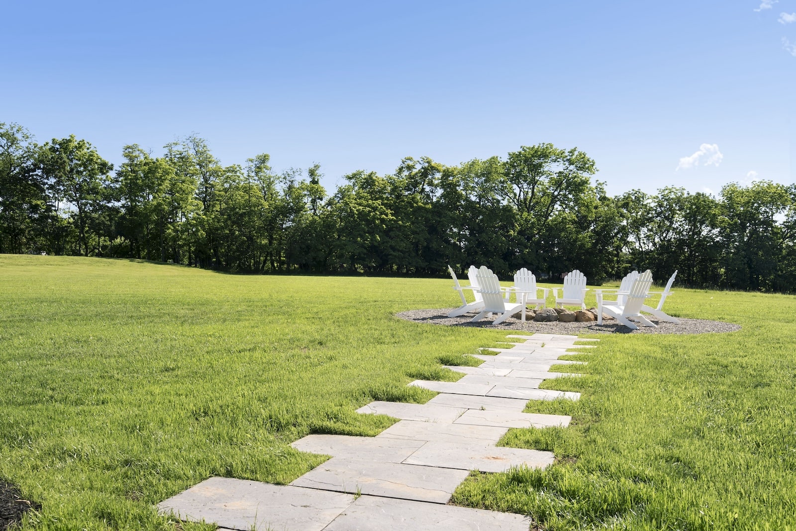 Stone path leading to a circular outdoor seating area with white Adirondack chairs arranged around a fire pit, set in a large grassy yard with trees in the background under a clear blue sky.