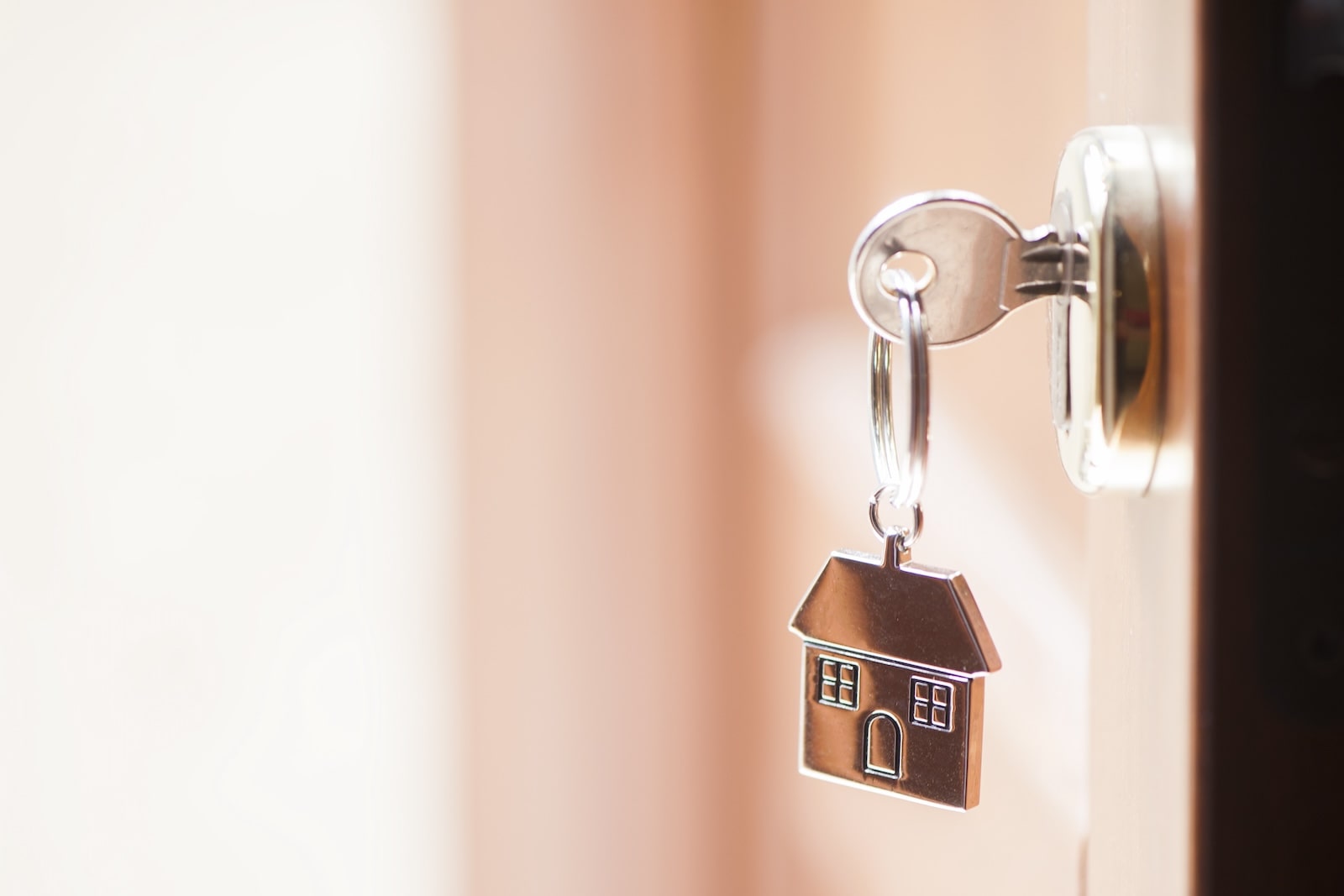 A close-up of a key inserted into a door lock, with a house-shaped keychain hanging from the key. The background is softly lit, suggesting natural daylight coming through the doorway.