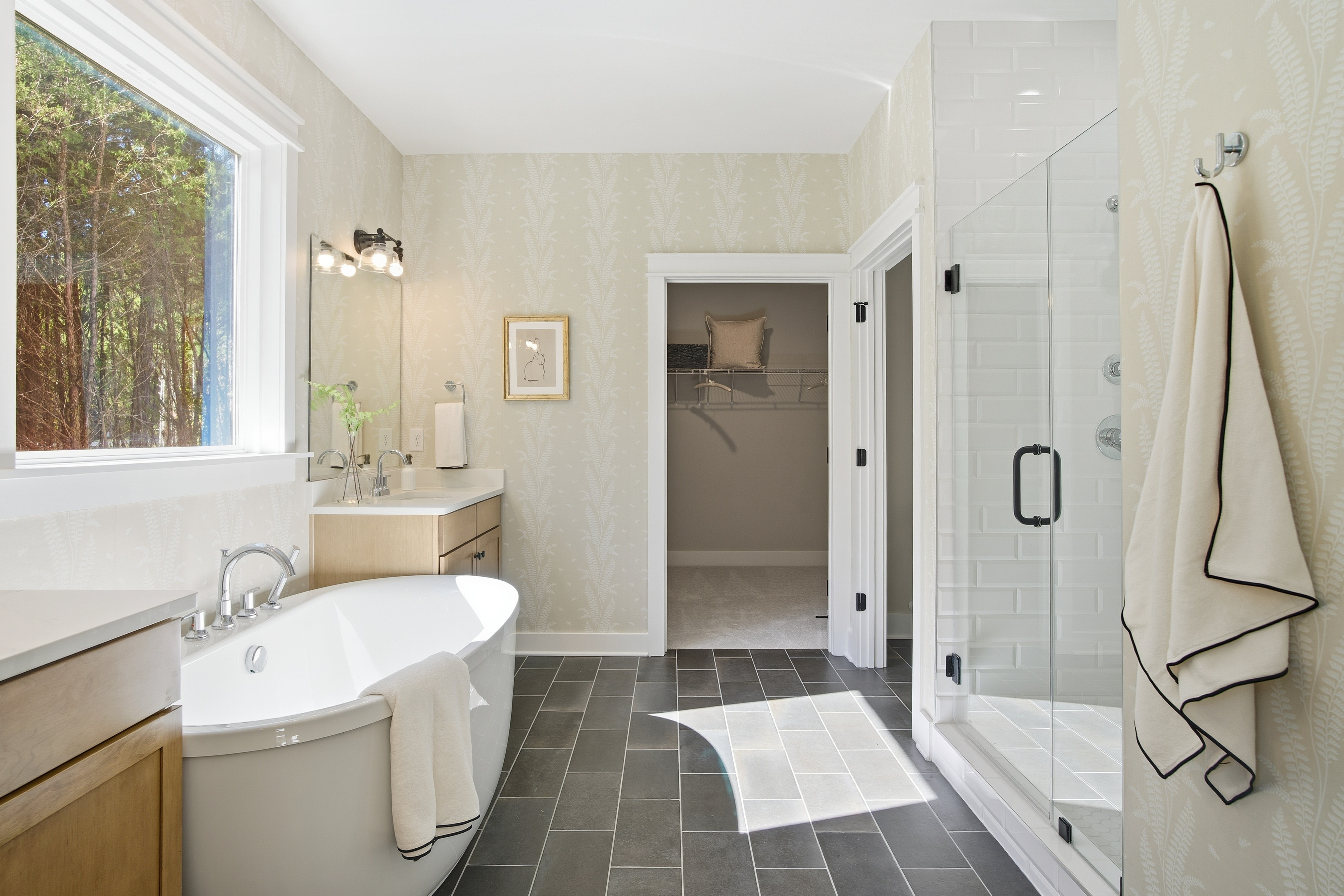 The bathroom from from the Appomattox plan from StyleCraft Homes. There's a tub, dark tile floors, ad shower, a sink and trees outside a large, wide window.