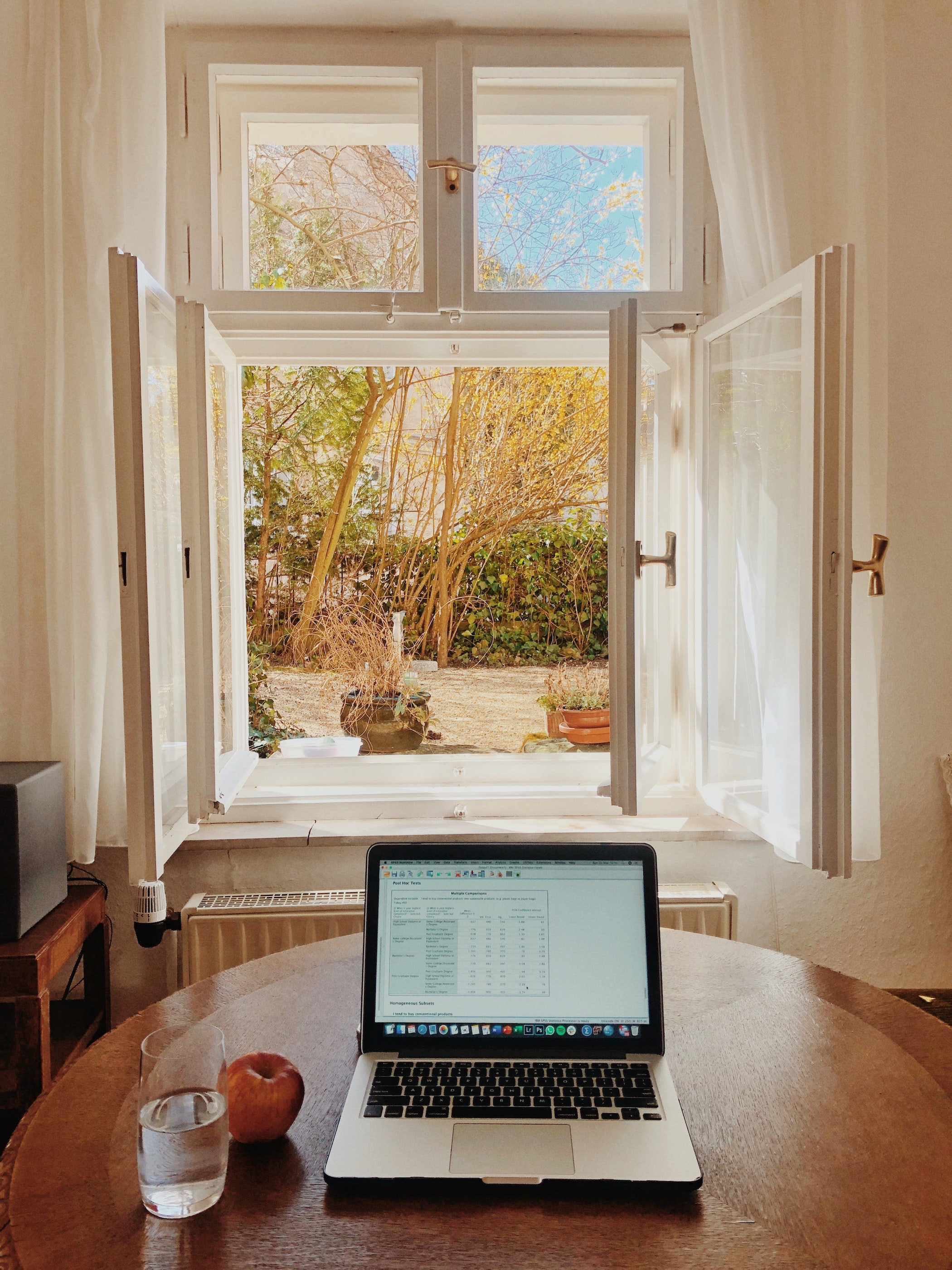 Image features a laptop open to a spreadsheet sitting on a kitchen table next to an apple and a glass of water, with an open window in the background opening to a foliage-filled yard.