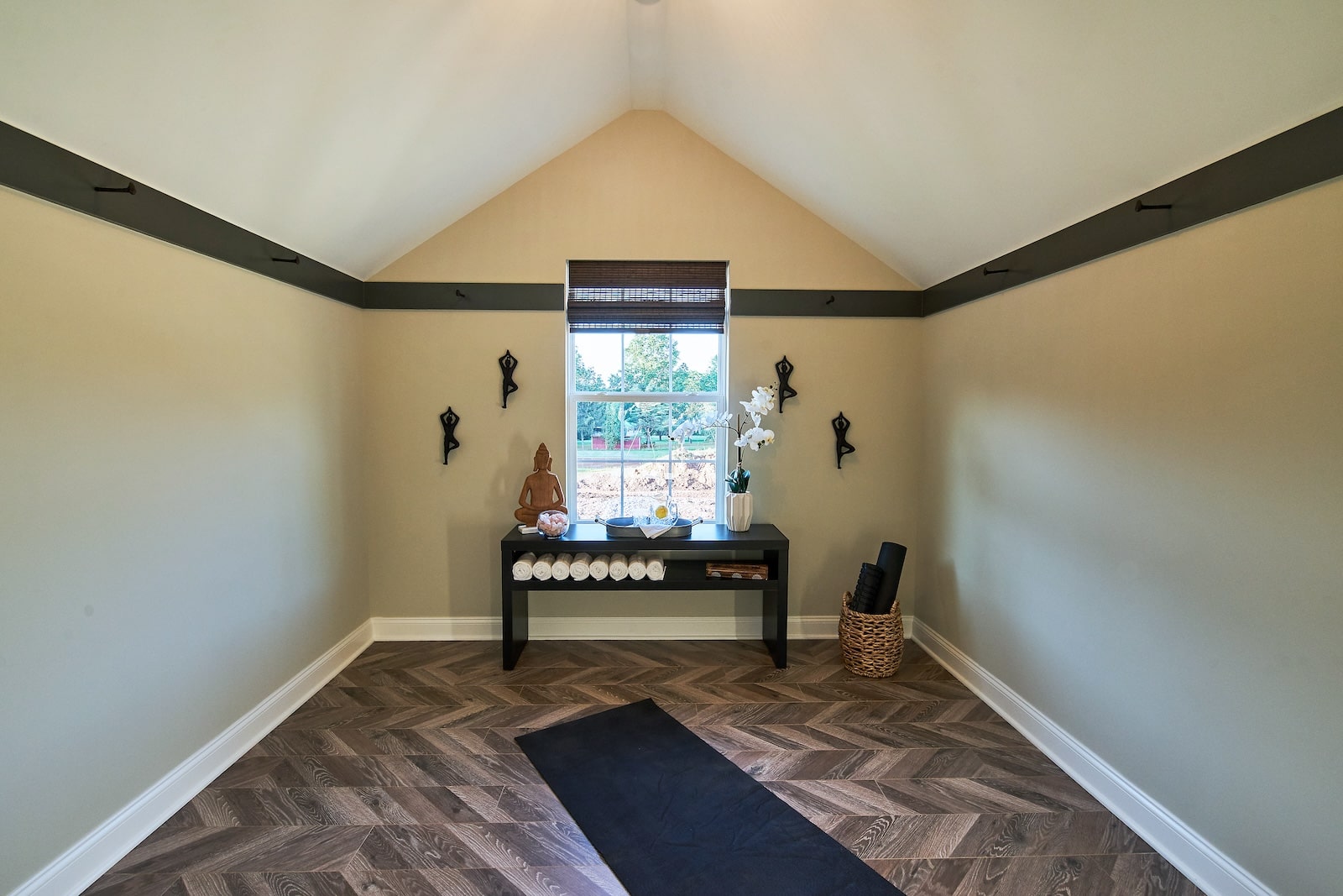 Peaceful home yoga room with a vaulted ceiling, wood-look herringbone floor, a single window, black bench with rolled towels and decor, and a yoga mat laid out on the floor.