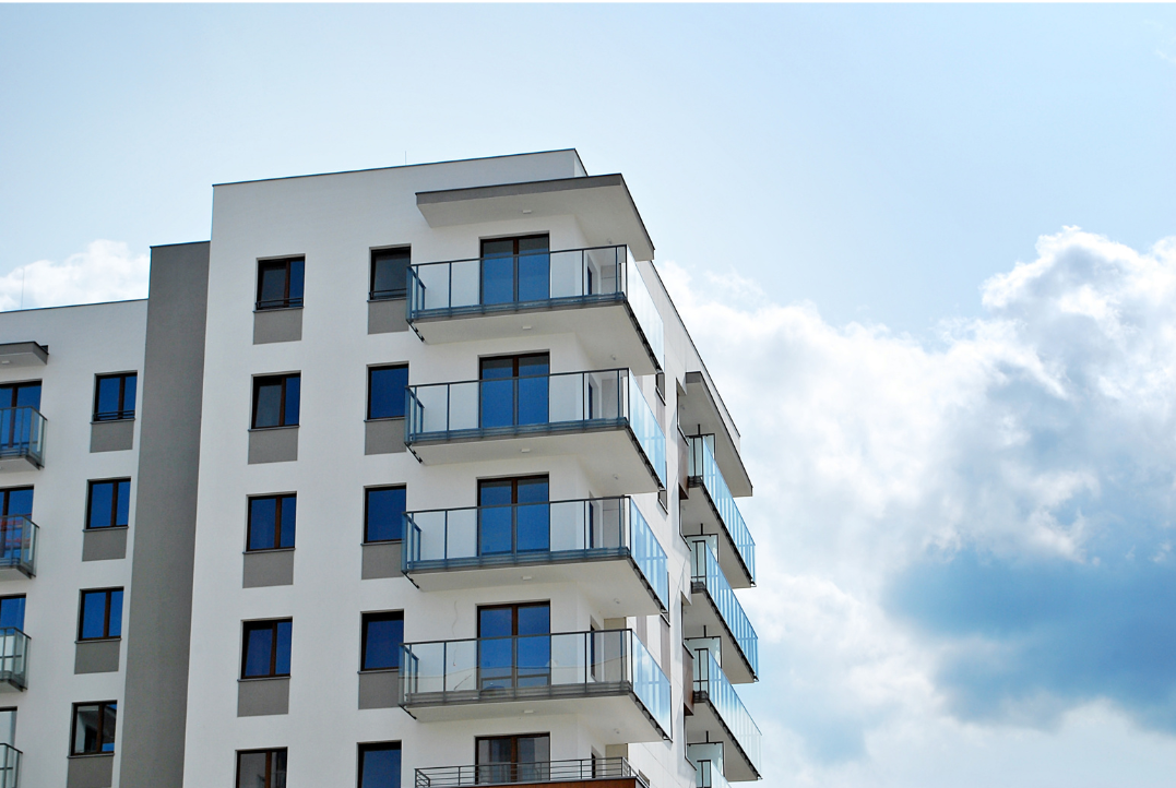 An apartment building with balconies. The sky in the background is light blue with some clouds.