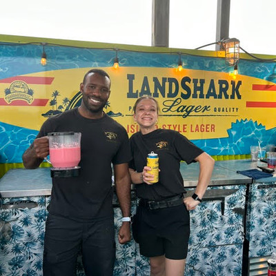 Two smiling staff members posing behind a LandShark Lager bar holding tropical drinks