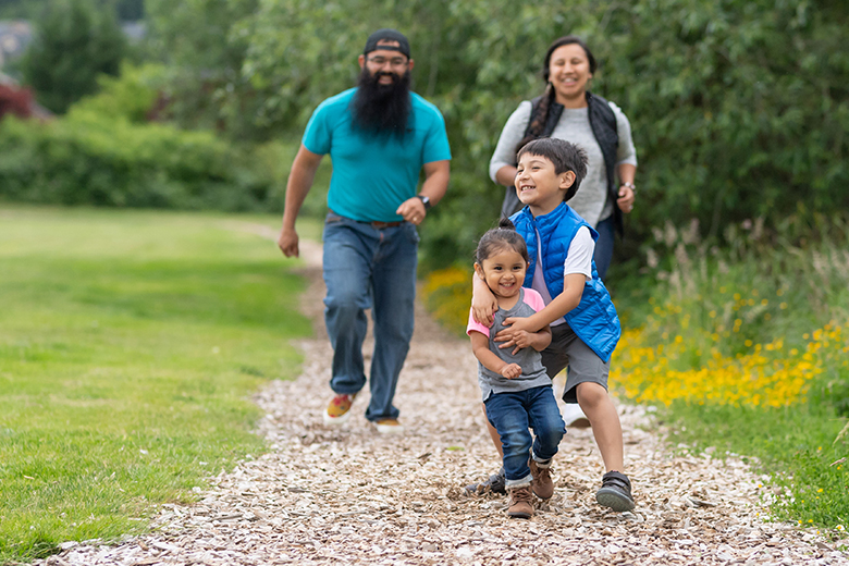 An elementary-age boy and his little sister run along a path out in nature. They're ahead of mom and dad and have big smiles. Big brother is playfully tackling his little sis.
