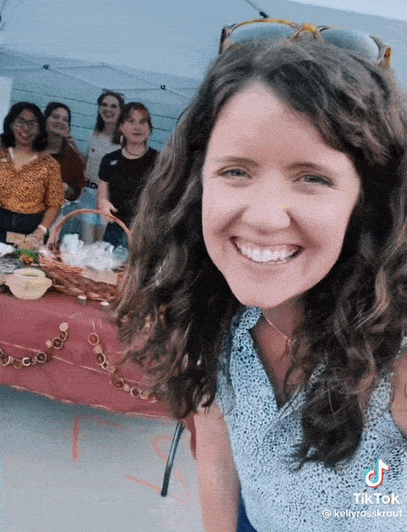 GIF of a TikTok posted by @KellyRossKout. In the video, Kelly is smiling and waving in front of a table with seven people who are also smiling and waving at the camera. There is white text with a green background overlaying the video that says, “Big Gay Market” with a rainbow emoji.