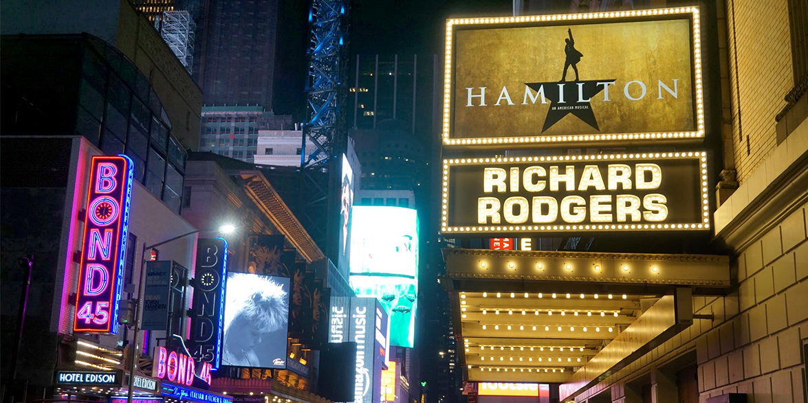 In New York City's Broadway District, a brightly lit, Richard Rogers Theatre marquee displays "Hamilton: An American Musical"