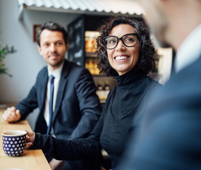 A smiling woman and man seated at a table engaged in discussion with another man.