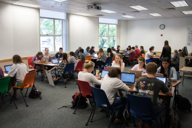 A classroom filled with students seated at grouped tables, working on laptops and papers during a class session, while an instructor stands at the front.