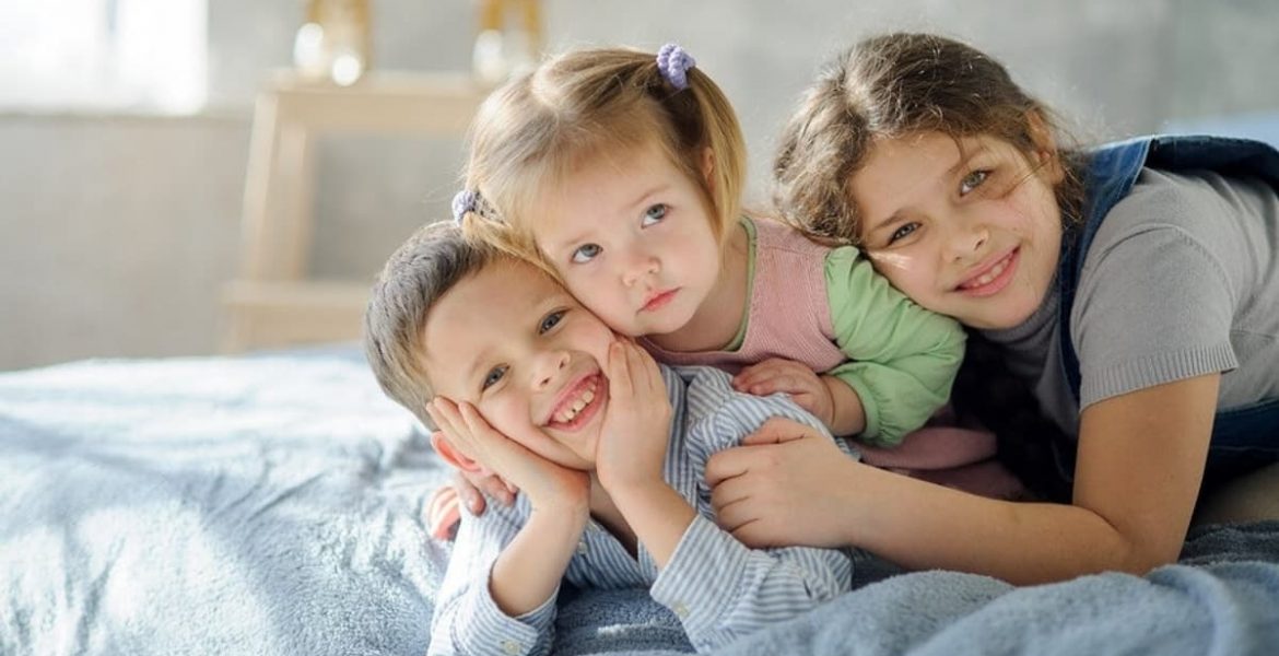 Fotografía de tres niños abrazados sobre una cama, reflejando la ternura, cercanía y complicidad entre hermanos.