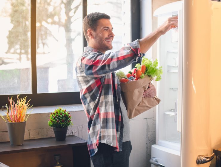 Hombre sonriente guardando verduras frescas en el refrigerador de su cocina en Estados Unidos.