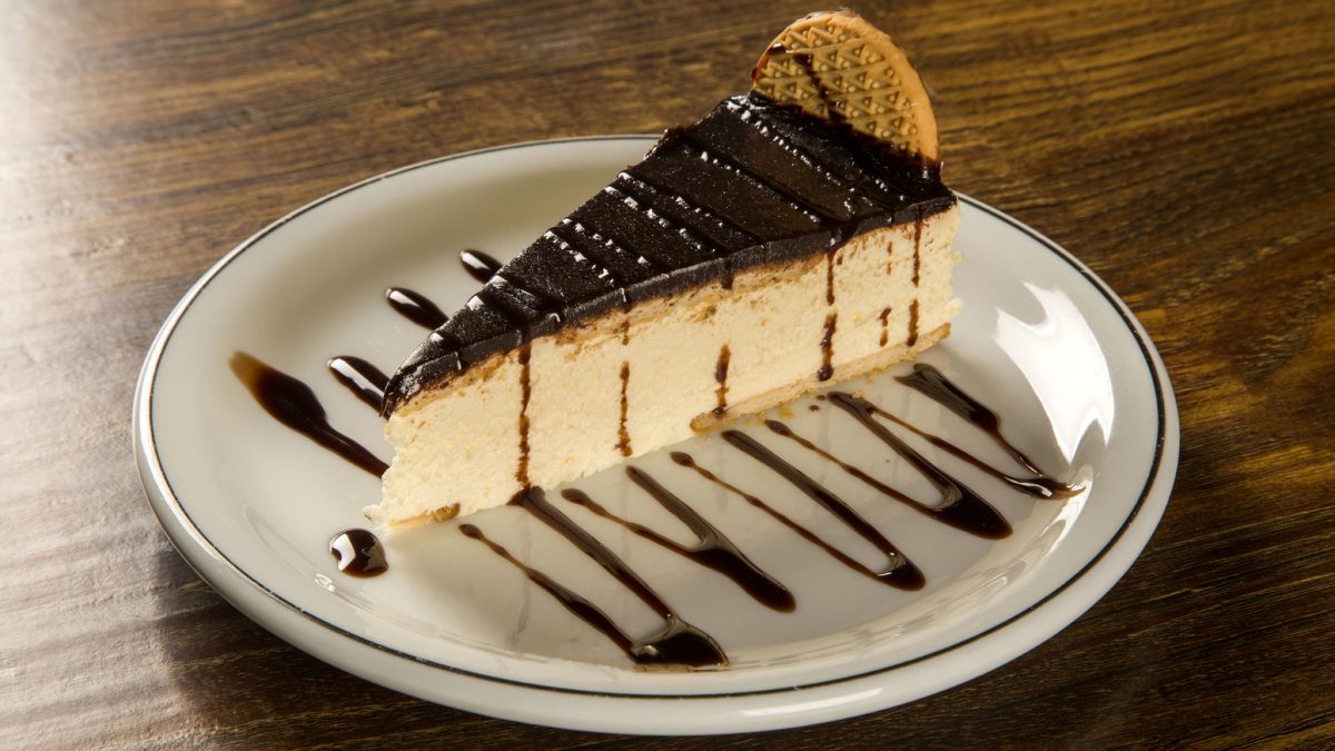 Rebanada de torta de queso cremosa con una brillante cobertura de chocolate, servida en un plato blanco y decorada con sirope de chocolate y una galleta crujiente.