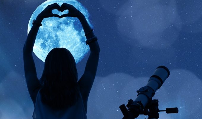 Mujer contemplando la luna llena con un telescopio, formando un corazón con las manos.