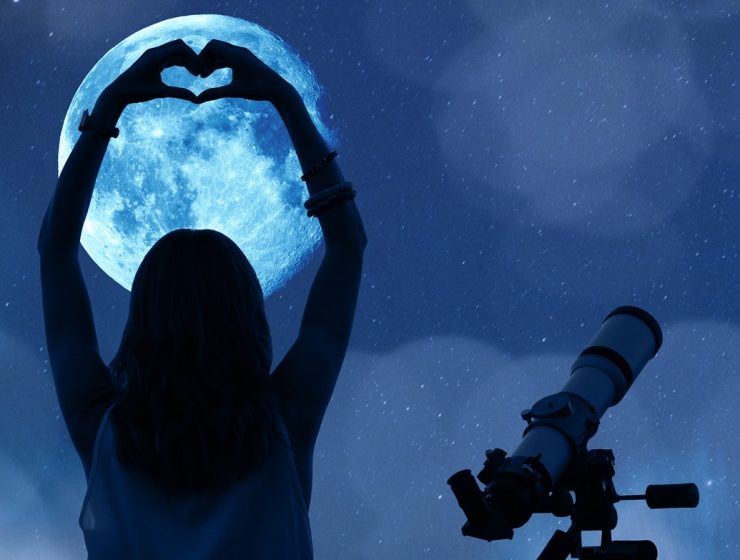 Mujer contemplando la luna llena con un telescopio, formando un corazón con las manos.
