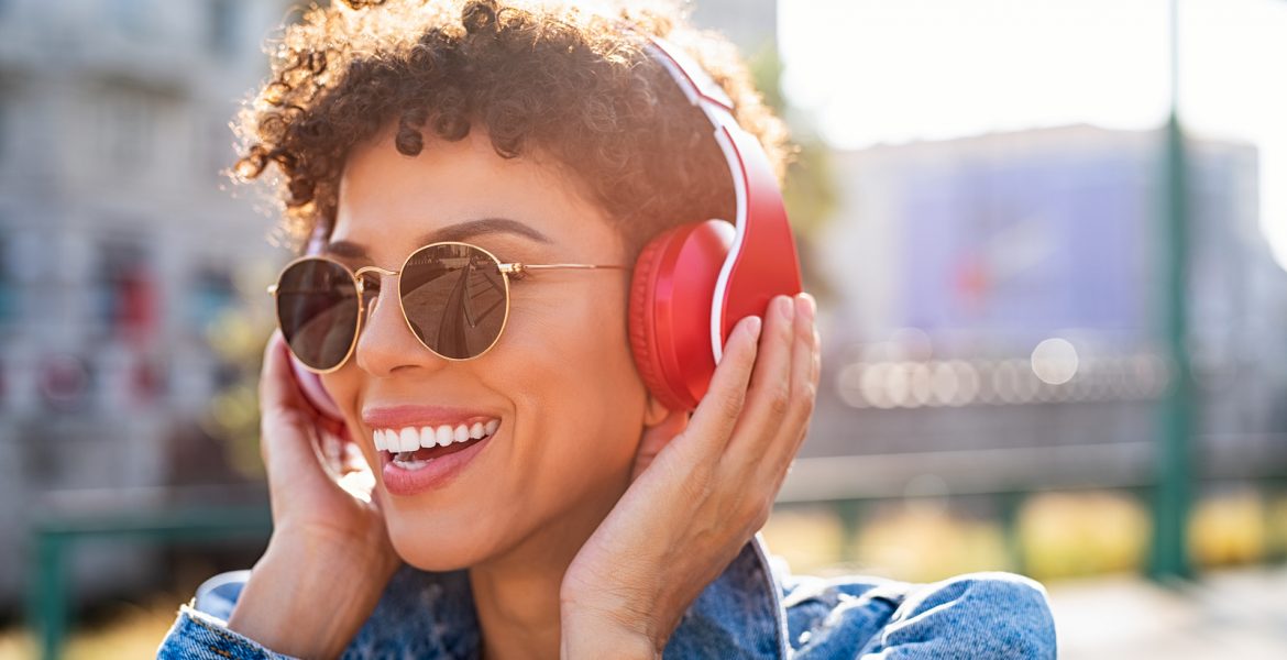 Mujer joven escuchando música con auriculares inalámbricos rojos al aire libre, sonriendo bajo el sol.