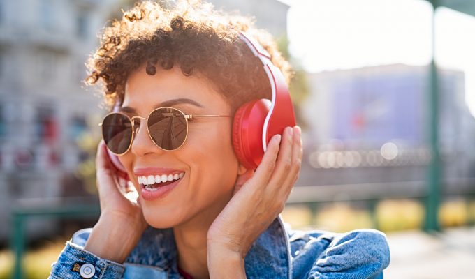 Mujer joven escuchando música con auriculares inalámbricos rojos al aire libre, sonriendo bajo el sol.