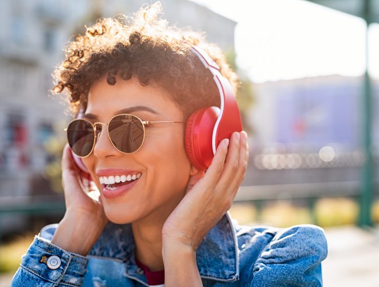 Mujer joven escuchando música con auriculares inalámbricos rojos al aire libre, sonriendo bajo el sol.