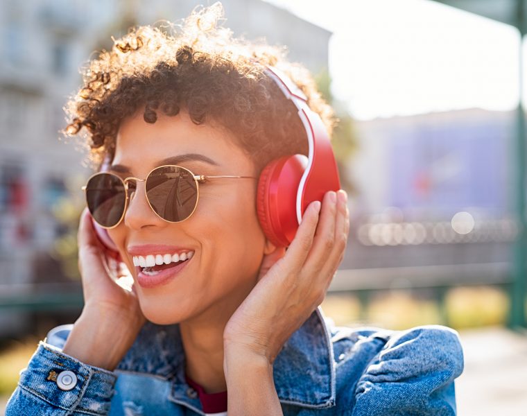 Mujer joven escuchando música con auriculares inalámbricos rojos al aire libre, sonriendo bajo el sol.