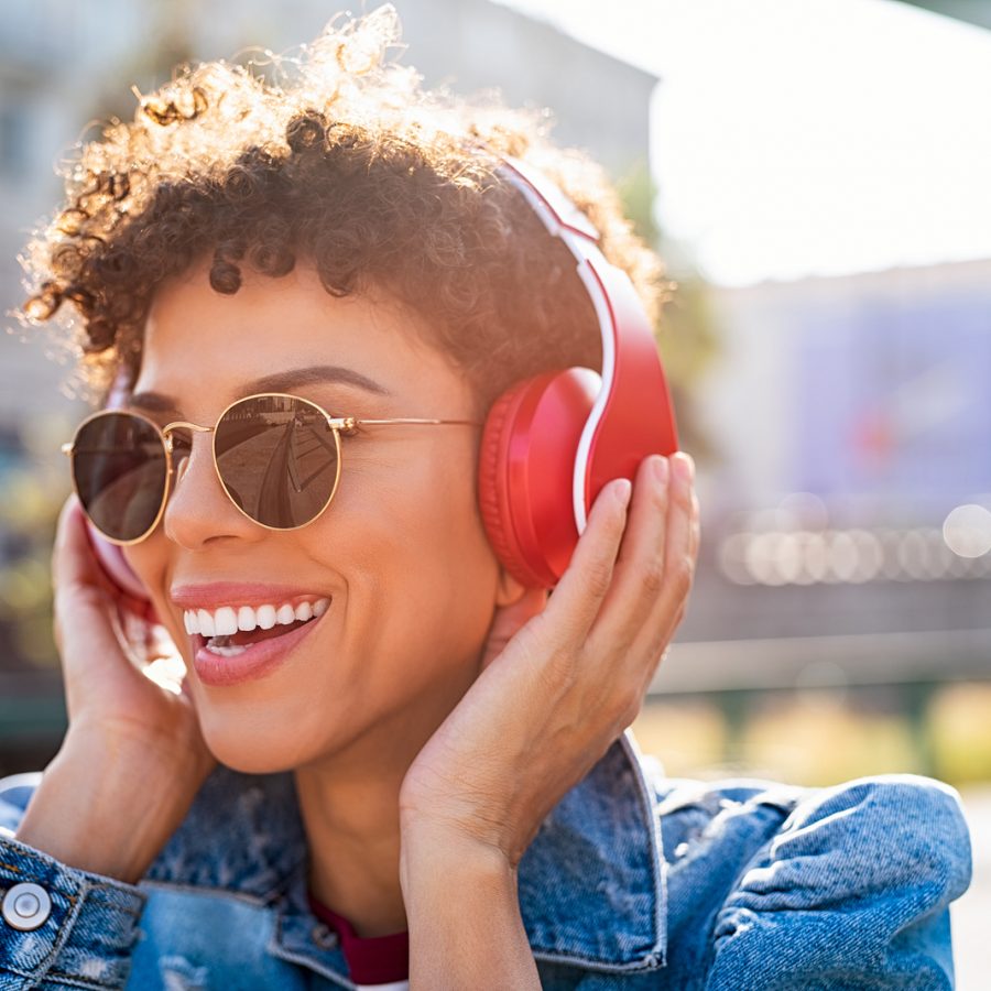 Mujer joven escuchando música con auriculares inalámbricos rojos al aire libre, sonriendo bajo el sol.