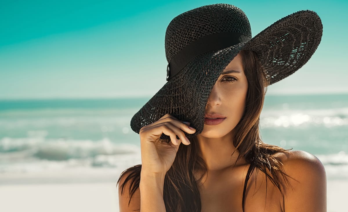 Mujer en la playa protegiéndose del sol con un elegante sombrero negro de ala ancha.