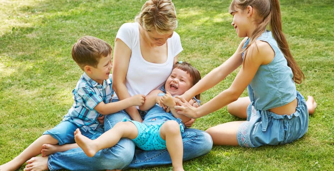 Madre jugando en el césped con sus tres hijos, símbolo de unión familiar y amor entre hermanos.