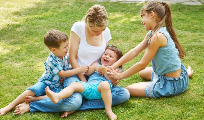 Madre jugando en el césped con sus tres hijos, símbolo de unión familiar y amor entre hermanos.