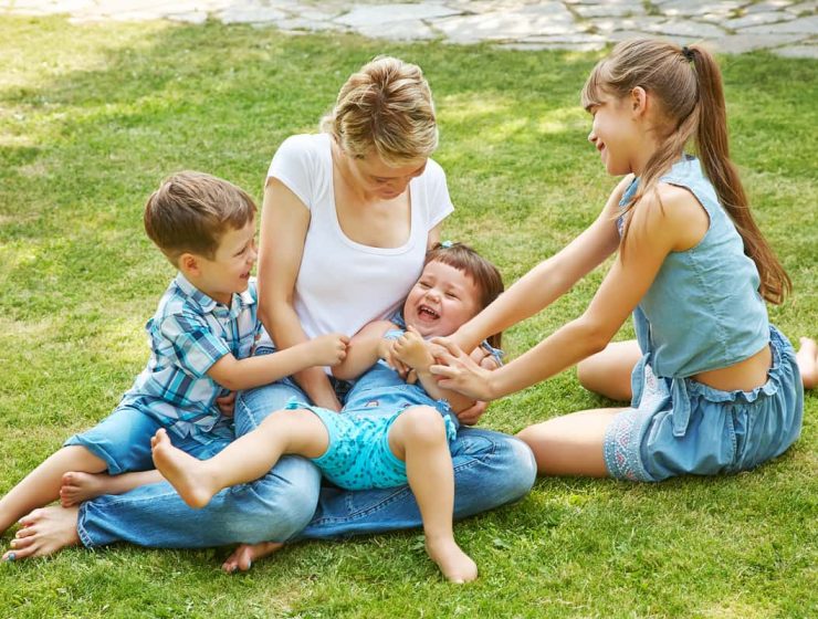 Madre jugando en el césped con sus tres hijos, símbolo de unión familiar y amor entre hermanos.
