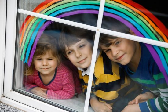 Arcoíris en la ventana: la mirada esperanzada de los niños durante el encierro. Tres niños mirando por la ventana con un arcoíris pintado, símbolo de esperanza.