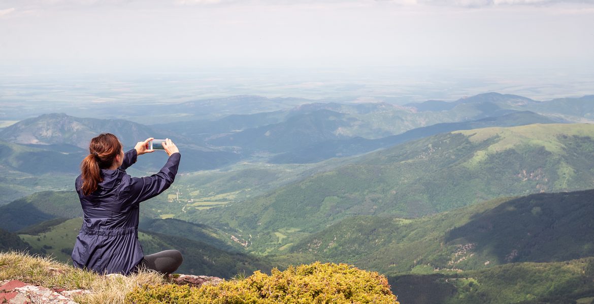 Mujer tomando una foto del paisaje montañoso con su celular desde la cima de una montaña.