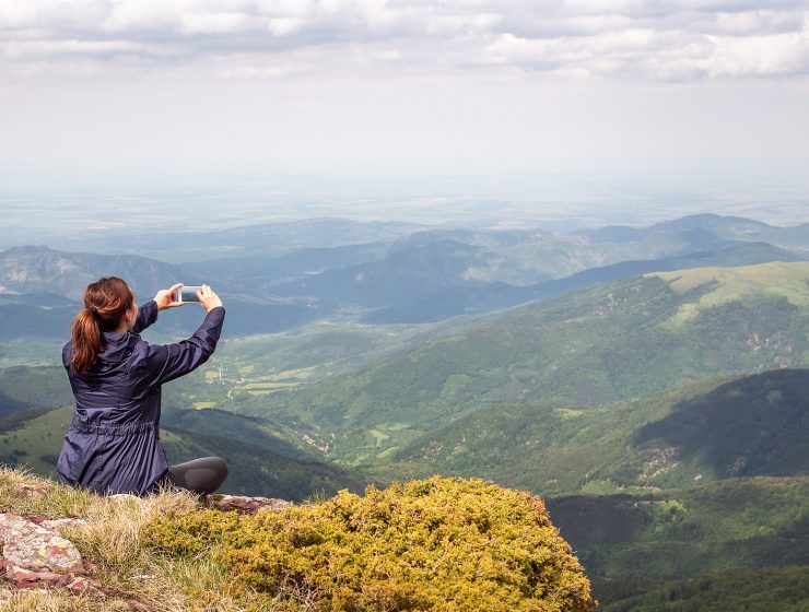 Mujer tomando una foto del paisaje montañoso con su celular desde la cima de una montaña.
