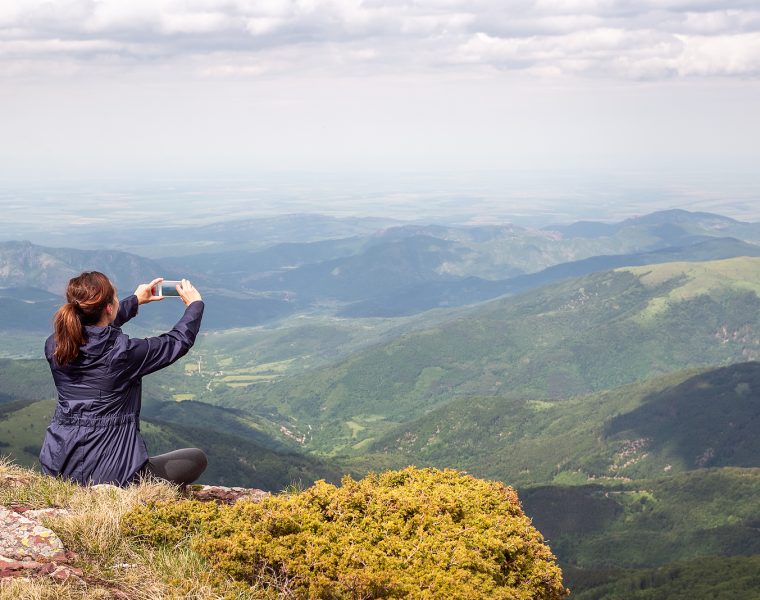 Mujer tomando una foto del paisaje montañoso con su celular desde la cima de una montaña.