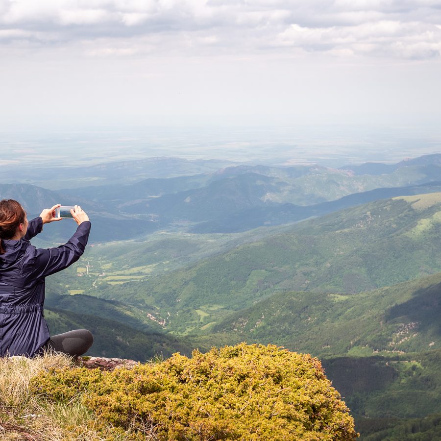 Mujer tomando una foto del paisaje montañoso con su celular desde la cima de una montaña.