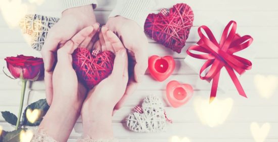 Manos sosteniendo un corazón rojo rodeado de velas, rosas y un regalo, símbolo de amabilidad y amor.