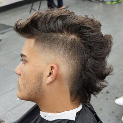 Hombre joven con un corte de pelo mullet moderno, caracterizado por un degradado (fade) muy marcado en los laterales y la nuca, contrastando con el cabello largo y texturizado en la parte superior y trasera.
