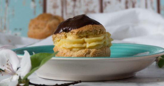 Profiterol relleno con crema pastelera y cubierto con chocolate, sobre un plato turquesa y fondo desenfocado.