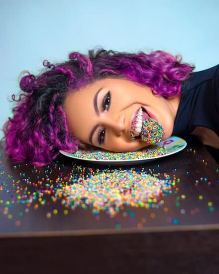 Con cabello morado, sacando la lengua cubierta de chispas de colores o sprinkles.