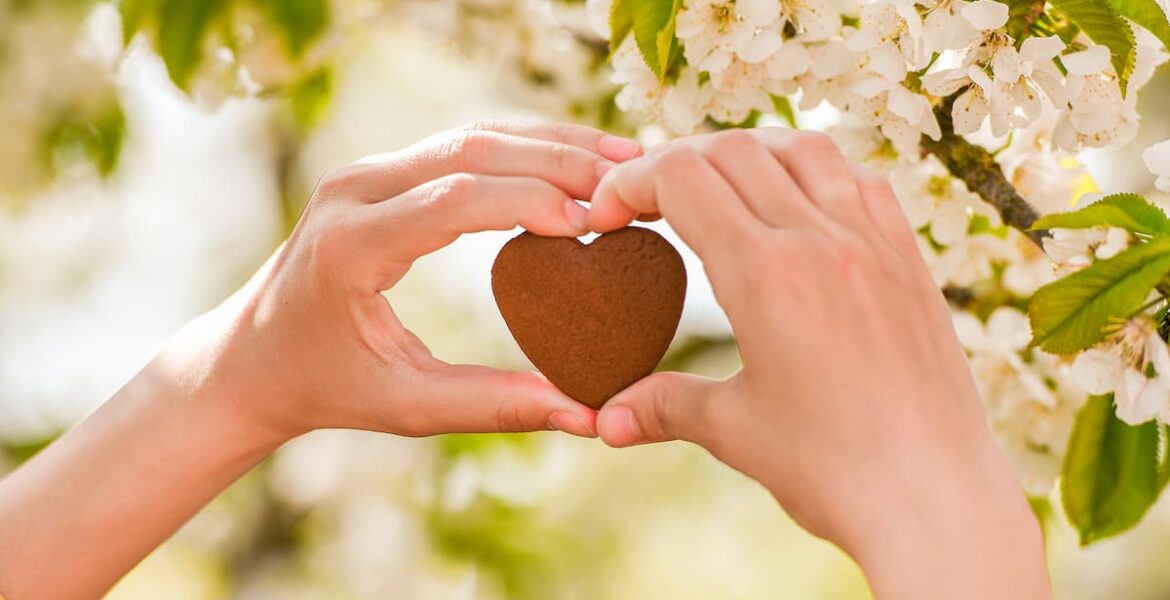 Manos formando un corazón con una galleta en forma de corazón frente a un árbol en flor, simbolizando la amabilidad.