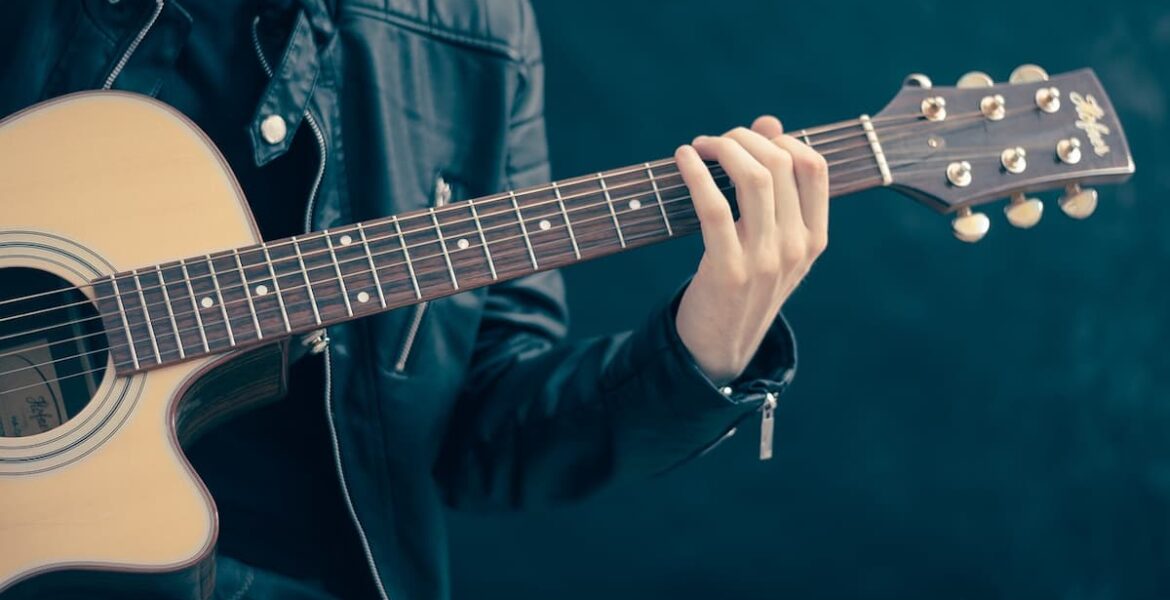 Un hombre practica guitarra mientras sigue una clase en línea desde una tablet, usando una app pensada para principiantes.