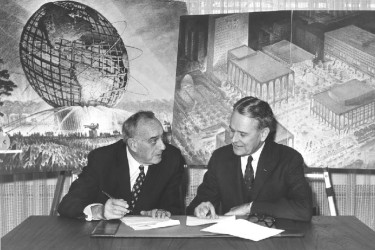 Two men sign documents in front of plans for the 1939 World's Fair.