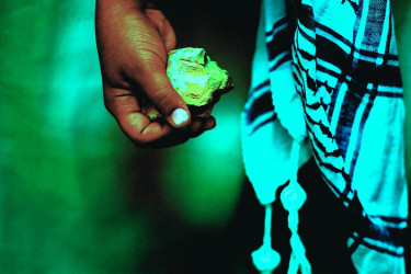 A hand holds a small rock, with a keffiyeh draping beside it.
