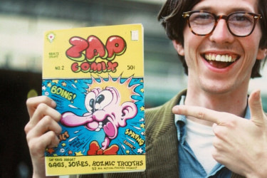 Robert Crumb holding up a cartoon book and pointing to it.