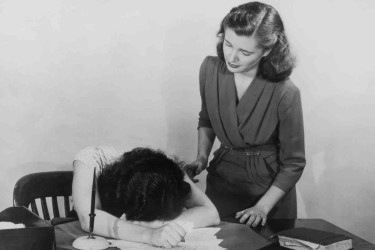 A woman comforting another woman, who has her face planted in a pile of papers.