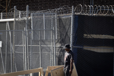 Photograph of a man carrying a wood board by a barbed wire fence.