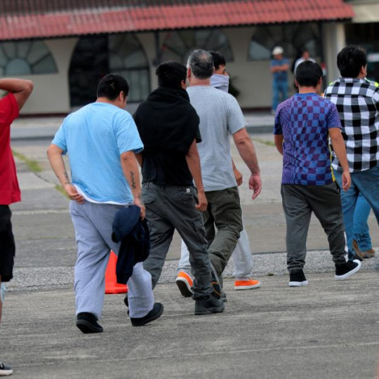 Deportees walk in a line while coordinators stand nearby in reflective yellow vests.