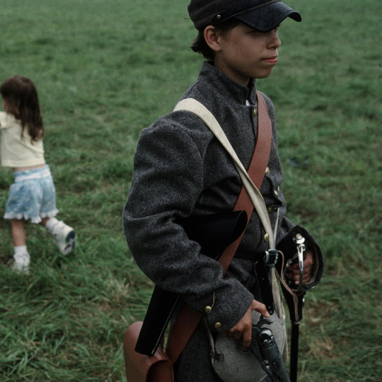 Image of a young boy carrying a pistol with women and children in the background.