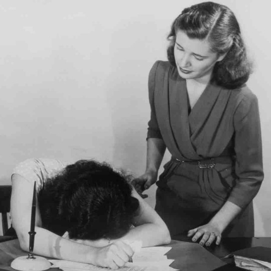 A woman comforting another woman, who has her face planted in a pile of papers.