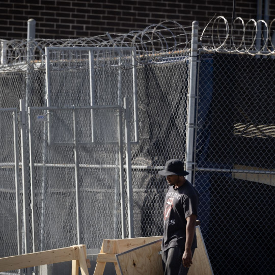 Photograph of a man carrying a wood board by a barbed wire fence.