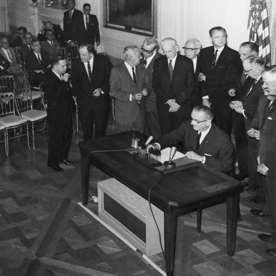 President Lyndon B. Johnson signs the Tonkin Gulf Resolution, in a room full of people.