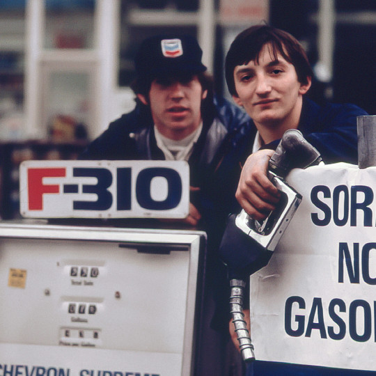 Service station attendants at a pump, with a sign announcing they are out of gasoline.