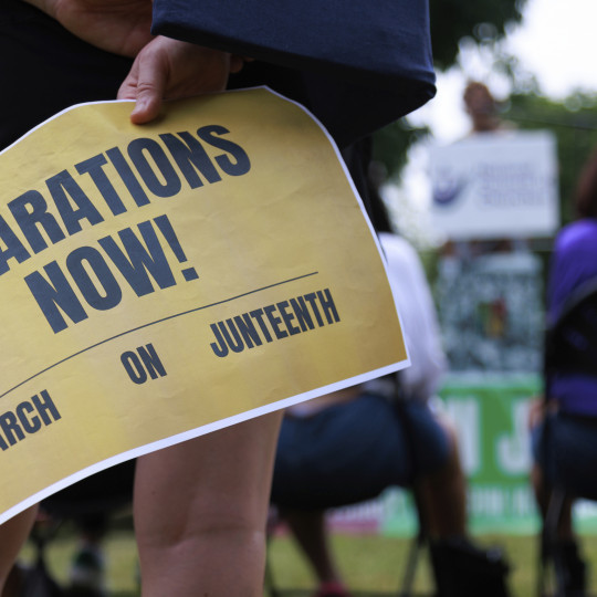 A person holds a sign reading “reparations now!”