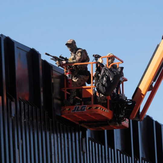 A man in fatigues holding a rifle while in a aerial lift platform truck to peek over the border wall.